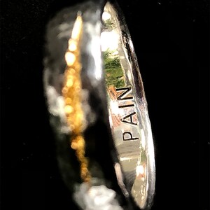Puede incluir: Un anillo de plata con una banda de oro y la palabra "PAIN" grabada en el lateral.