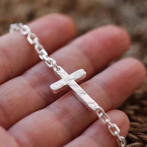 May include: A silver-colored bracelet featuring a textured cross pendant. The cross is connected to a chain-link bracelet. The bracelet is made of a shiny, metallic material.