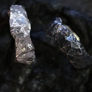 May include: Two textured metal rings, one silver and one dark gray, are displayed against a dark background. The rings have a rough, uneven surface, suggesting a handcrafted or organic design. The close-up shot highlights the details of the metalwork.