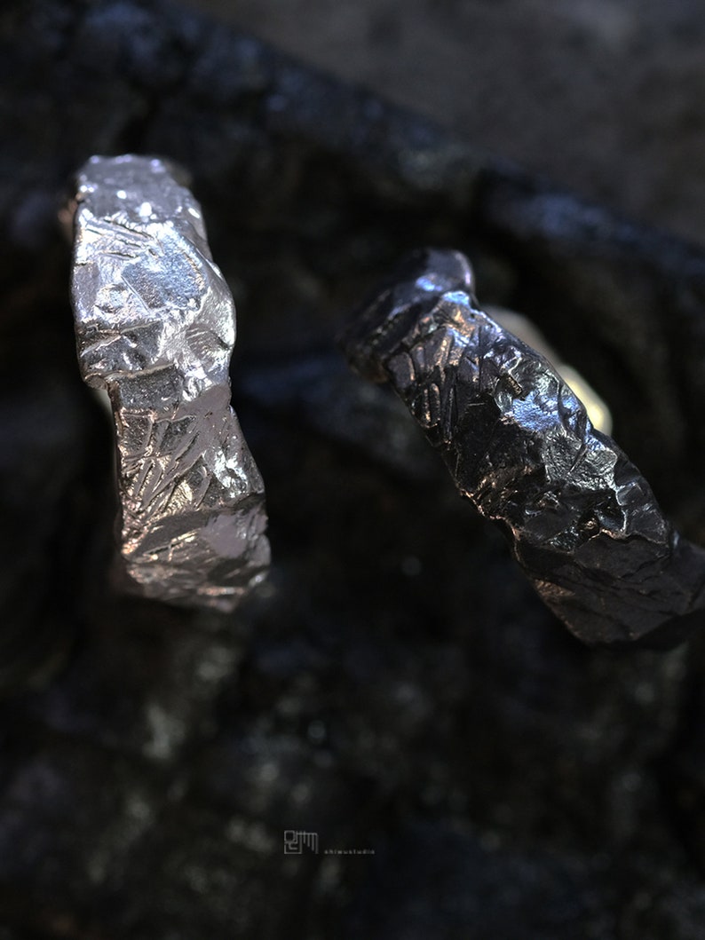 May include: Two textured metal rings, one silver and one dark gray, are displayed against a dark background. The rings have a rough, uneven surface, suggesting a handcrafted or organic design. The close-up shot highlights the details of the metalwork.