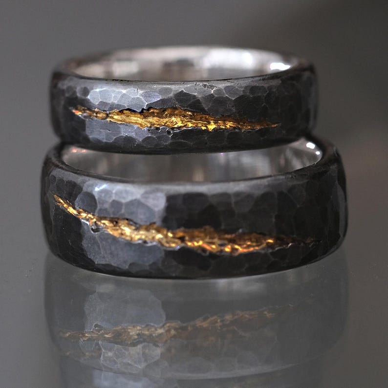 Puede incluir: Dos alianzas de boda de color negro y plata con una fina franja dorada que recorre cada alianza. Las alianzas tienen una textura martillada.