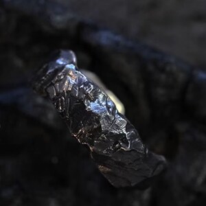 May include: A dark, textured ring with a rugged, organic appearance. The ring's surface has a rough, uneven texture, with a dark, metallic sheen. The design suggests a handcrafted or artisan-made piece, suitable for everyday wear.