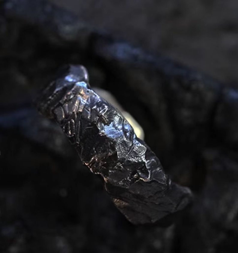 May include: A dark, textured ring with a rugged, organic appearance. The ring's surface has a rough, uneven texture, with a dark, metallic sheen. The design suggests a handcrafted or artisan-made piece, suitable for everyday wear.