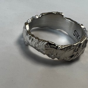 Puede incluir: Un anillo plateado con un diseño texturizado y orgánico. El anillo tiene una superficie rugosa e irregular, y el interior es liso. La palabra "SIKO" está grabada en el interior del anillo.