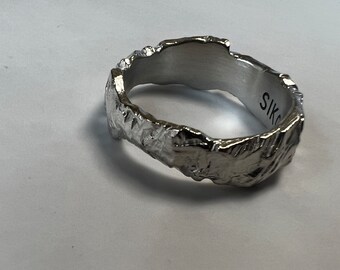 May include: A silver-colored ring with a textured, organic design. The ring has a rough, uneven surface, and the interior is smooth. The word "SIKO" is engraved on the inside of the ring.