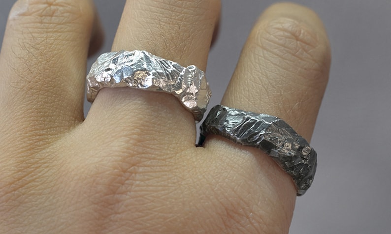 May include: Two textured silver rings are displayed on a hand. One ring is bright silver, the other is a darker, oxidized silver. The rings have a rugged, organic appearance, suggesting a handcrafted design. The rings are likely for everyday wear.