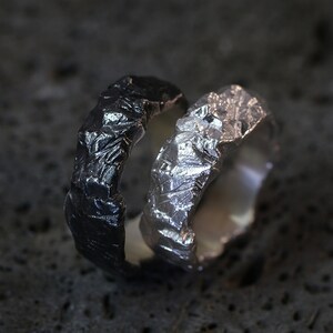 May include: Two textured rings, one black and one silver, are displayed. The rings have a rough, organic appearance, with a crinkled surface. The silver ring reflects light, while the black ring has a matte finish. The rings are likely made of metal.