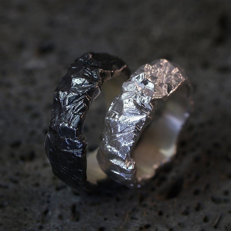May include: Two textured rings, one black and one silver, are displayed. The rings have a rough, organic appearance, with a crinkled surface. The silver ring reflects light, while the black ring has a matte finish. The rings are likely made of metal.
