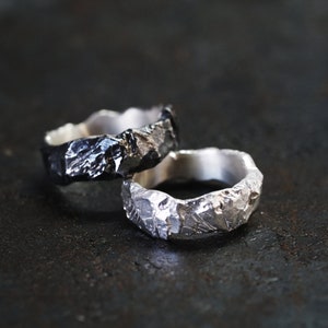 May include: Two textured metal rings, one dark and one silver, are displayed. The rings have a rugged, organic appearance, with irregular surfaces. The rings are likely made of sterling silver or a similar metal. The rings are a unique jewelry item.