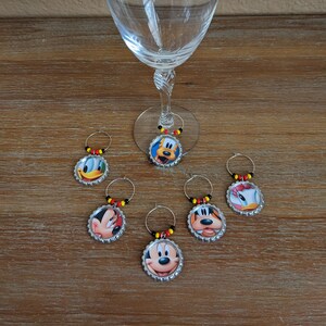 May include: Six wine glass charms featuring different Disney characters. The charms are silver with a clear plastic ring and black, yellow, and red beads. The characters include Mickey Mouse, Minnie Mouse, Donald Duck, Goofy, and Pluto.