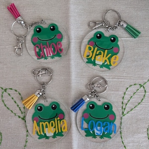 Puede incluir: Cuatro llaveros redondos de acrílico transparente con ilustraciones de ranas verdes y los nombres "Chloe", "Blake", "Amelia" y "Logan" en letras de colores. Cada llavero tiene un anillo partido plateado y un borla de colores.