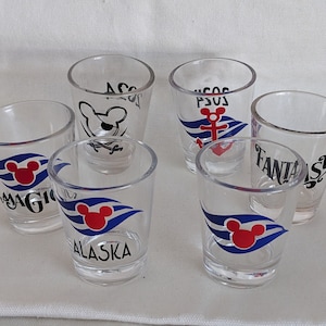 May include: Six clear glass shot glasses with different Disney cruise ship logos. The logos include "Magic", "Alaska", "Fantasy", "Dream", "Wonder", and an anchor with "SOS" above it.