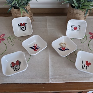 May include: Six white square ceramic trinket dishes with various Disney-themed designs. The designs include Mickey Mouse, Minnie Mouse, a heart with Mickey Mouse hands, an anchor with a Disney cruise line logo, and the word "LOVE" with a Mickey Mouse head.