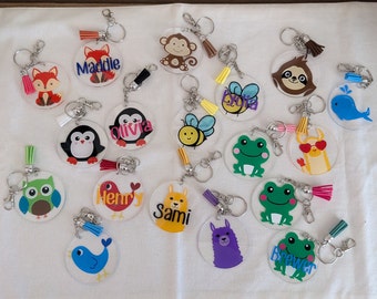 May include: A collection of 15 colorful, personalized keychains with various animal and insect designs. Each keychain features a clear acrylic circle with a colorful image and a name printed on it. The keychains are attached to silver key rings with colorful tassels.