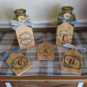 Peut inclure: Cinq petites planches à découper en bois avec des nœuds en ruban noir et blanc. Les planches sont décorées d'une couronne et d'une lettre au centre. Les lettres sont C, K, C et U. Les planches sont sur une surface à carreaux.