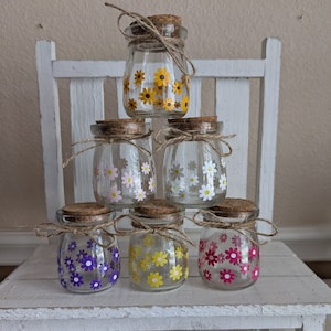 Peut inclure: Six bocaux en verre avec des couvercles en liège et de la ficelle attachée autour du cou. Chaque bocal a un motif floral différent avec des fleurs jaunes, blanches, violettes et roses.