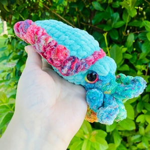 May include: A crocheted plush toy shaped like a cuttlefish with a blue body, pink and purple tentacles, and a single black eye. The toy is held in a hand against a green leafy background.