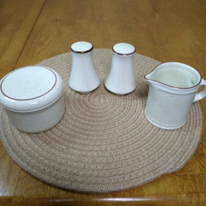 May include: A set of four ceramic kitchenware items in a beige and brown color scheme. The set includes a creamer, two salt and pepper shakers, and a small covered dish. The items are arranged on a woven placemat.