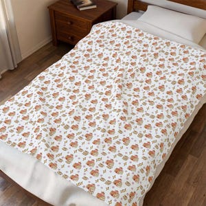 Op de afbeelding: Witte deken met een herhalend patroon van hartvormige pannenkoeken, sterren en geometrische vormen in oranje, bruin en roze tinten. De deken ligt op een bed met een wit kussen en een houten nachtkastje.