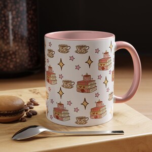 Puede incluir: Taza de cerámica blanca con interior y asa rosas. La taza presenta un patrón repetido de tazas de té, pilas de libros con velas y estrellas doradas. Una cuchara y un pastelito están en primer plano.