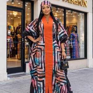 May include: A floor-length kaftan featuring a solid orange centre panel and a black, orange, and teal geometric pattern on the sides. The garment has wide sleeves and a matching headwrap. A small black handbag is held at the side of the dress.