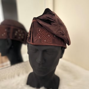 May include: A dark brown hat with an embroidered design and small, sparkling embellishments. The hat is displayed on a black mannequin head, with a reflection in a mirror. The hat has a triangular shape and a soft, textured appearance.