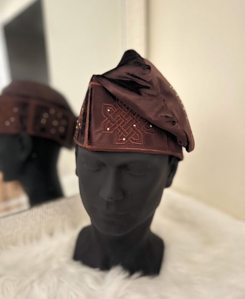 May include: A dark brown hat with an embroidered design and small, sparkling embellishments. The hat is displayed on a black mannequin head, with a reflection in a mirror. The hat has a triangular shape and a soft, textured appearance.