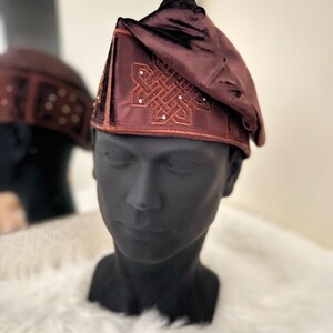 May include: A maroon velvet hat with an embroidered design and small, decorative embellishments. The hat is displayed on a black mannequin head, with a blurred reflection in the background. The hat has a unique, folded shape.