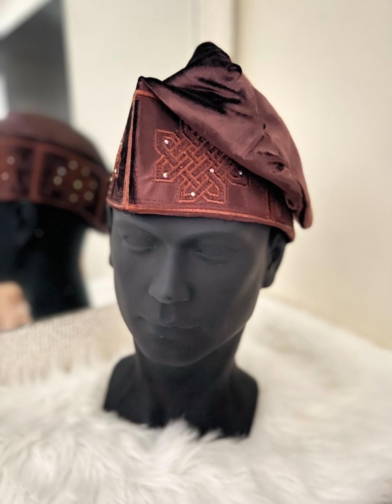May include: A maroon velvet hat with an embroidered design and small, decorative embellishments. The hat is displayed on a black mannequin head, with a blurred reflection in the background. The hat has a unique, folded shape.