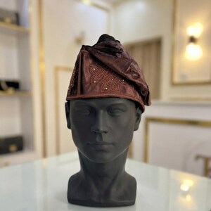 May include: A brown, ornate hat with a decorative pattern and small embellishments. The hat is displayed on a black mannequin head. The hat's design features a folded, draped style, and the fabric has a subtle sheen.