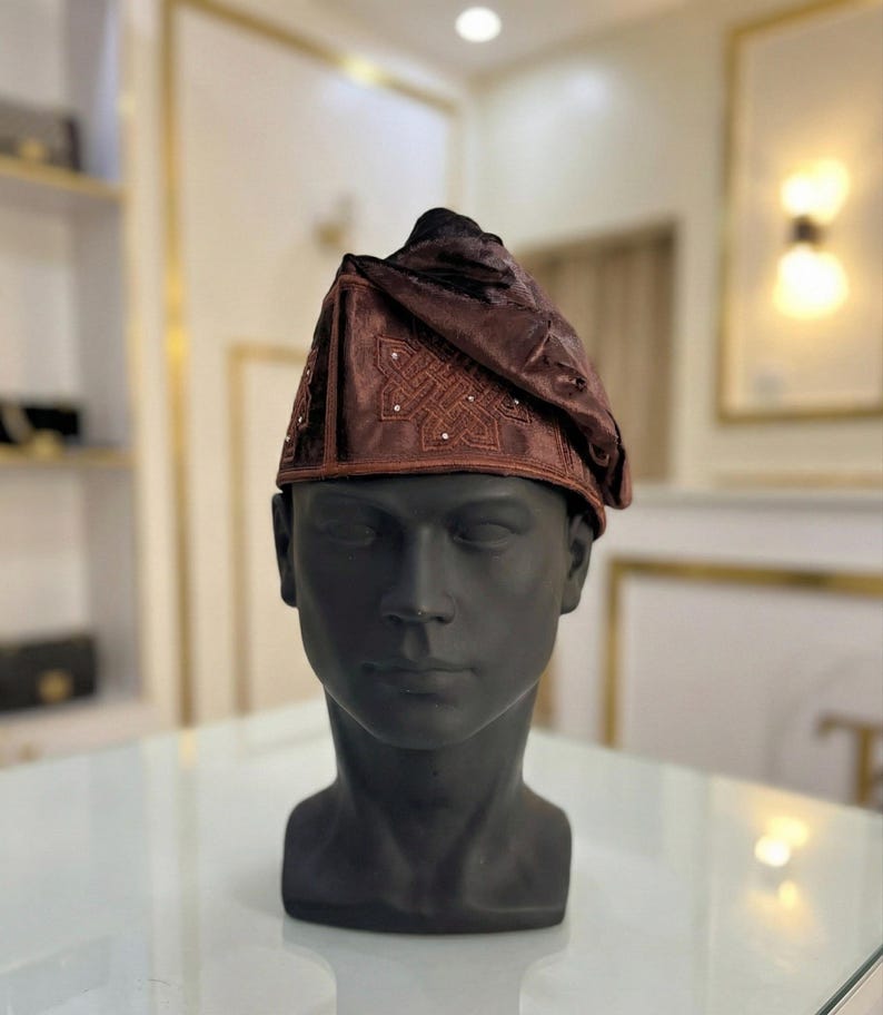May include: A brown, ornate hat with a decorative pattern and small embellishments. The hat is displayed on a black mannequin head. The hat's design features a folded, draped style, and the fabric has a subtle sheen.