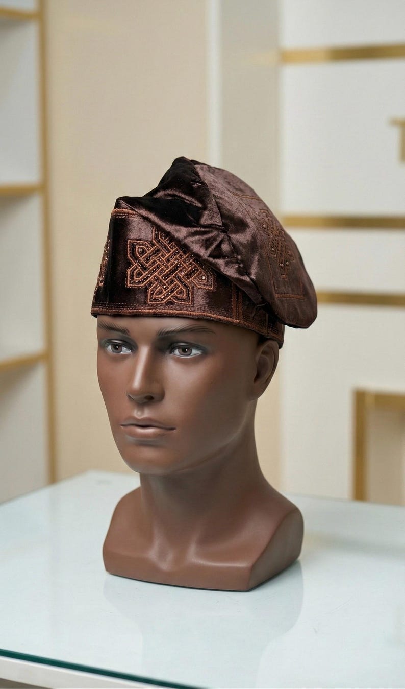 May include: A brown velvet hat with intricate embroidered patterns on the brim and crown, displayed on a mannequin head. The hat has a rounded shape with a slight peak, and the embroidery is in a lighter shade of brown.