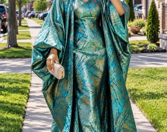 May include: A floor-length teal and gold metallic dress with a matching open-front cape featuring wide sleeves. The fabric has a textured, branch-like pattern throughout. A small, rectangular, gold-toned clutch is held in one hand.