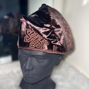May include: A brown velvet hat with an embroidered geometric design. The hat is displayed on a black mannequin head. The hat has a soft, textured appearance and a dark brown color.