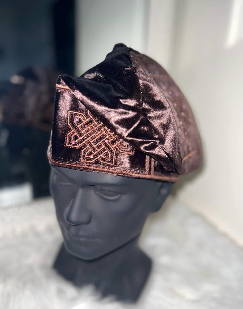 May include: A brown velvet hat with an embroidered geometric design. The hat is displayed on a black mannequin head. The hat has a soft, textured appearance and a dark brown color.