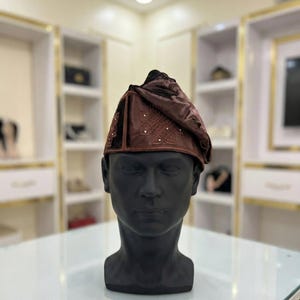May include: A brown velvet cap with intricate embroidery and small, sparkling embellishments. The cap is displayed on a black mannequin head, showcasing its design and texture. The hat is a traditional style.