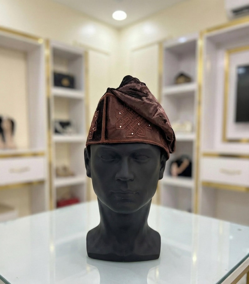 May include: A brown velvet cap with intricate embroidery and small, sparkling embellishments. The cap is displayed on a black mannequin head, showcasing its design and texture. The hat is a traditional style.