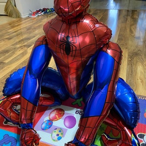 K&ouml;nnte beinhalten: Ein gro&szlig;er roter und blauer aufblasbarer Spiderman-Ballon mit einem wei&szlig;en Spinnenlogo auf der Brust. Der Ballon liegt auf einer bunten Spielmatte.