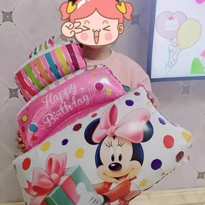 May include: A child wearing a sparkly crown holds a stack of colorful foil balloons. The balloons are shaped like a cake and feature the words "Happy Birthday!" and a cartoon image of Minnie Mouse.