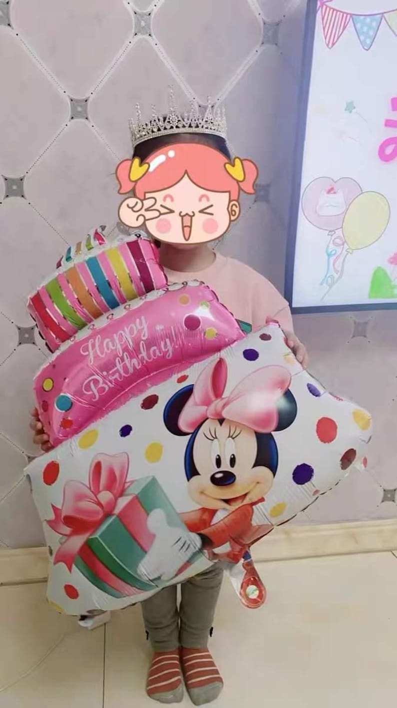 May include: A child wearing a sparkly crown holds a stack of colorful foil balloons. The balloons are shaped like a cake and feature the words "Happy Birthday!" and a cartoon image of Minnie Mouse.