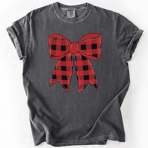 May include: Dark gray t-shirt featuring a large red and black buffalo plaid bow graphic. The bow is centered on the shirt and has a classic, festive design. The shirt has a vintage, worn look.