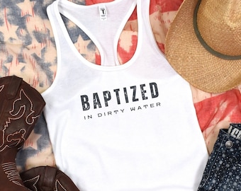 May include: A white tank top with the text "BAPTIZED IN DIRTY WATER" is displayed with brown cowboy boots, denim shorts, and a straw cowboy hat. The items are arranged on a red, white, and blue patterned scarf.