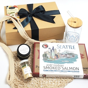 May include: A gift basket filled with various items, including a brown box with a black ribbon, a reusable mesh bag, a glass jar with matches, a small tin of candle, a glass jar with a bamboo lid and a straw, and a box with a Seattle skyline illustration and the text "Wild Sockeye Smoked Salmon".