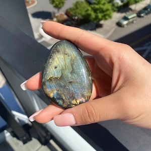 May include: A smooth, oval-shaped labradorite gemstone with iridescent blue, green, and gold hues. The stone is held in a hand.