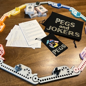 Pode incluir: Um tabuleiro de jogo com uma pista circular feita de peças de plástico coloridas. O tabuleiro é cercado por um baralho de cartas, uma bolsa preta com o texto "Pegs and Jokers", e uma bolsa preta menor com o texto "Pegs".