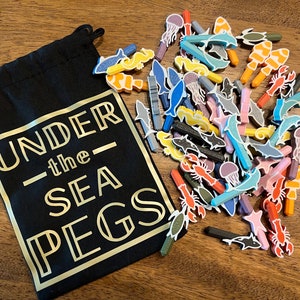 Pode incluir: Uma bolsa de cordão preta com o texto "UNDER the SEA PEGS" em letras douradas. A bolsa está cheia de pinos de madeira de criaturas marinhas coloridas, incluindo peixes, medusas, cavalos-marinhos e lagostas.