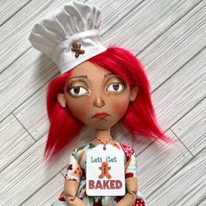 Puede incluir: Muñeca hecha a mano con cabello rojo y gorro de chef, adornada con un hombre de jengibre. La muñeca lleva un vestido y sostiene una etiqueta que dice "Lets Get Baked" con un gráfico de hombre de jengibre. La muñeca tiene una expresión ligeramente triste.