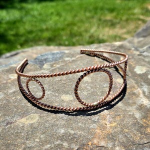 May include: A copper-colored cuff bracelet with a twisted rope design. The bracelet features two circular accents and a simple, open design. The jewelry is displayed on a stone surface with a blurred green background.