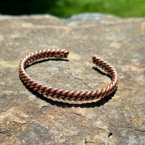 May include: A copper-colored, twisted wire cuff bracelet. The bracelet is open-ended and sits on a textured, gray-brown stone surface. The twisted design gives the bracelet a braided appearance.