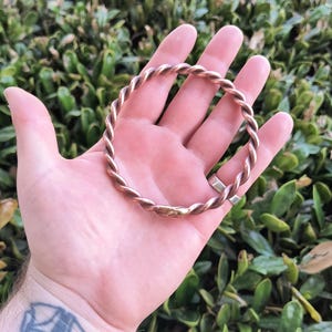 May include: A twisted copper bracelet, with a reddish-brown color, is held in a hand against a green leafy background.